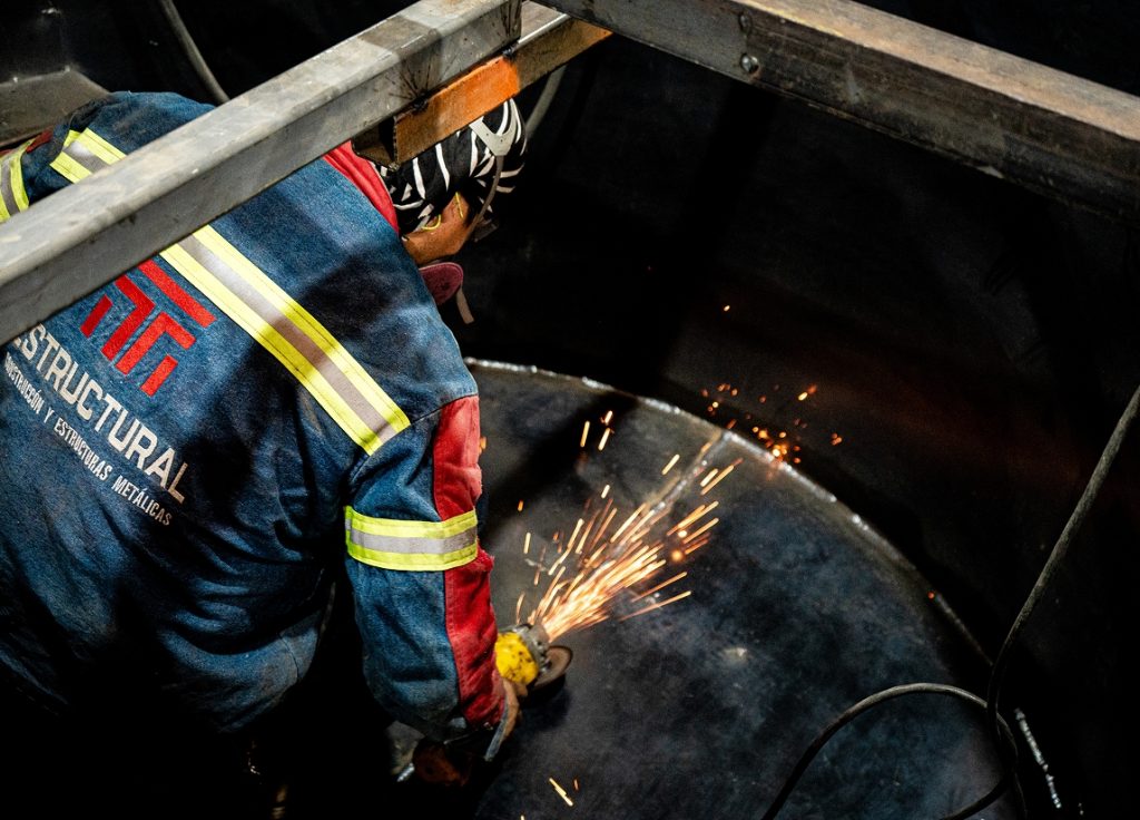 Técnico soldador con uniforme de seguridad TM ESTRUCTURAL trabajando sobre tanque metálico en proceso de fabricación.